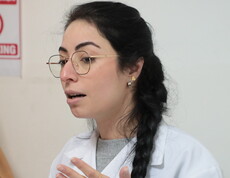 Frau mit Brille und geflochtenem Zopf in weißem Kittel spricht und gestikuliert.