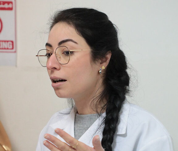 Frau mit Brille und geflochtenem Zopf in weißem Kittel spricht und gestikuliert.