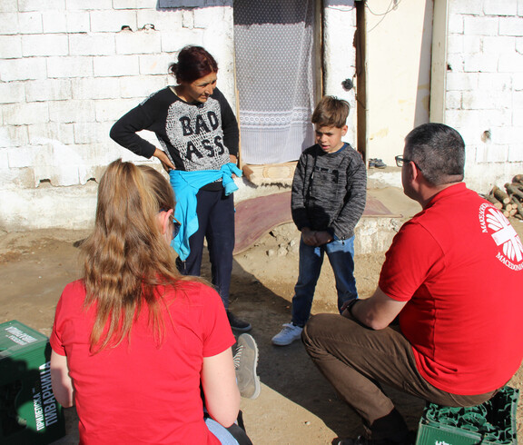 Caritas-Projektreferentin Theresa Sacher steht mit einem Jungen und einer Frau vor einem einfachen Haus. Zwei weitere Erwachsene sitzen oder stehen gegenüber, während ein Gespräch stattfindet.