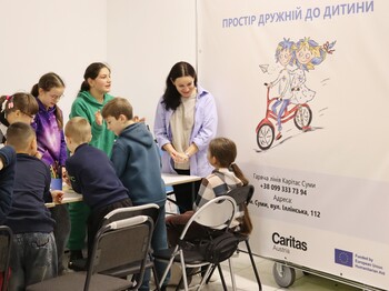 Mehrere Kinder sitzen mit einer Betreuerin an einem Tisch und malen; im Hintergrund ein Plakat zum EU-Projekt „Protection“ in der Ost-Ukraine.