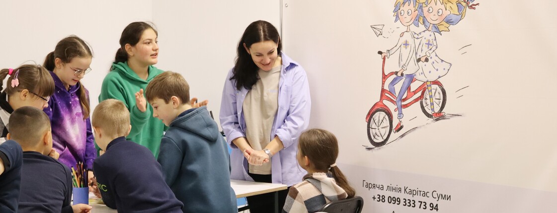 Mehrere Kinder sitzen mit einer Betreuerin an einem Tisch und malen; im Hintergrund ein Plakat zum EU-Projekt „Protection“ in der Ost-Ukraine.