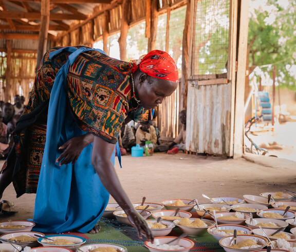 eine südsudanesische Frau beugt sich zu gefüllten Tellern, die am Boden stehen.