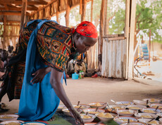 eine südsudanesische Frau beugt sich zu gefüllten Tellern, die am Boden stehen.