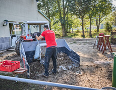 Ein freiwilliger Helfer schaufelt Schlamm in eine Traktorschaufel vor einem Gebäude in Hofstetten-Grünau. Der Mann trägt ein rotes T-Shirt mit der Aufschrift „Wir helfen“. Im Hintergrund sind Bäume und Werkzeuge zu sehen, die für die Aufräumarbeiten verwendet werden.
