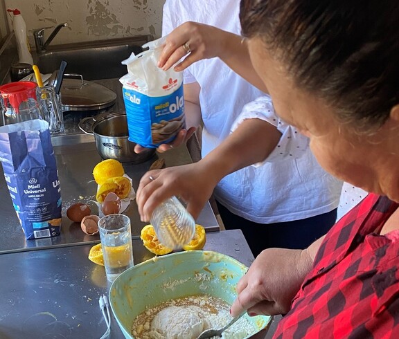 Zwei Frauen stehen in einer Küche und mischen gemeinsam Teig in einer Schüssel; auf der Arbeitsfläche liegen verschiedene Zutaten.