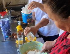 Zwei Frauen stehen in einer Küche und mischen gemeinsam Teig in einer Schüssel; auf der Arbeitsfläche liegen verschiedene Zutaten.