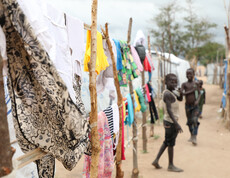 Zwei Kinder laufen zwischen provisorischen Unterkünften im Camp Gumbo, im Vordergrund hängen bunte Kleidungsstücke an Holzpfählen.