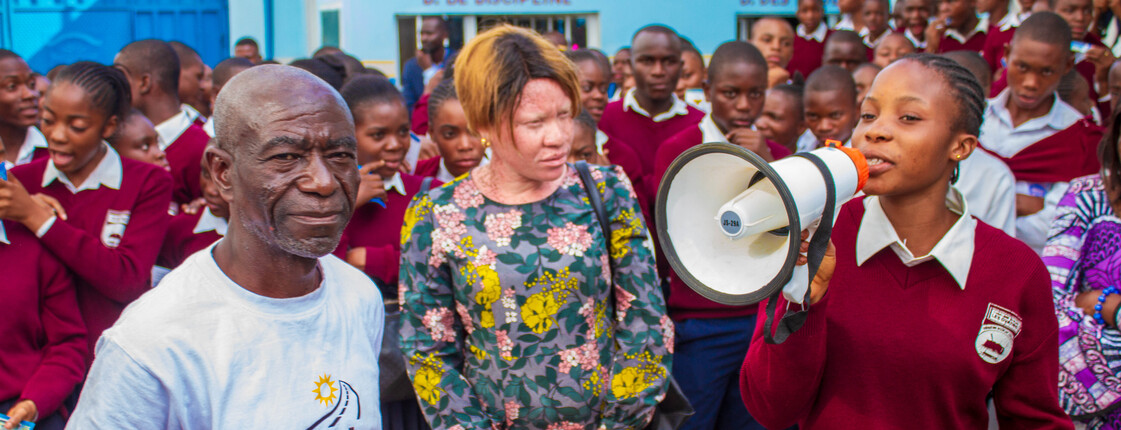 Eine große Gruppe von Schülerinnen und Schülern steht im Schulhof. Im Vordergrund spricht ein Mädchen in Schuluniform durch ein Megafon. Neben ihr stehen ein älterer Mann im FLEM-T-Shirt und eine Frau in einem geblümten Kleid. Alle blicken aufmerksam in Richtung der Sprecherin.