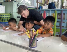 Ein junger Erwachsener unterstützt mehrere Kinder beim Schreiben und Lernen an einem Tisch im Klassenzimmer.