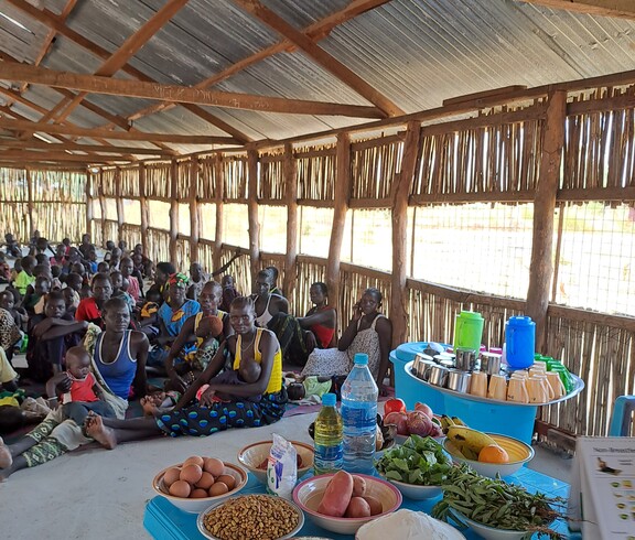 eine Gruppe südsudanesischer Mütter sitzt vor einem blauen Tisch, der mit verschiedenen Zutaten bedeckt ist