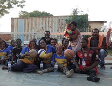 Gruppe von Sitzvolleyballspieler*innen sitzt lächelnd auf dem Spielfeld und hält Volleybälle in der Hand, im Hintergrund ein Netz und Zuschauende.