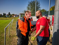 Ein Mann in einer orangen Arbeitsjacke schüttelt lächelnd die Hand eines Freiwilligen in einem roten Caritas-T-Shirt. Im Hintergrund ist ein Traktor und ein Sportplatz zu sehen, wo weitere Helfer an den Aufräumarbeiten beteiligt sind.