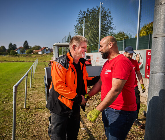 Ein Mann in einer orangen Arbeitsjacke schüttelt lächelnd die Hand eines Freiwilligen in einem roten Caritas-T-Shirt. Im Hintergrund ist ein Traktor und ein Sportplatz zu sehen, wo weitere Helfer an den Aufräumarbeiten beteiligt sind.