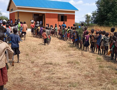 Viele Schulkinder stehen in einer langen Reihe vor einem kleinen Gebäude mit blauem Dach und warten auf ihre Mahlzeit.