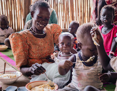 Eine südsudanesische Mutter und ihr Baby sitzen mit anderen Kindern am Boden und essen.