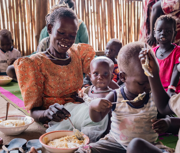 Eine südsudanesische Mutter und ihr Baby sitzen mit anderen Kindern am Boden und essen.
