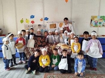 In einem armenischen Kindergarten stehen eine Frau und viele Kinder, die weiße Kochschürzen umgebunden haben, zu einem Gruppenfoto zusammen.