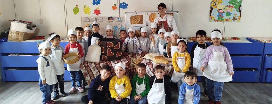 In einem armenischen Kindergarten stehen eine Frau und viele Kinder, die weiße Kochschürzen umgebunden haben, zu einem Gruppenfoto zusammen.