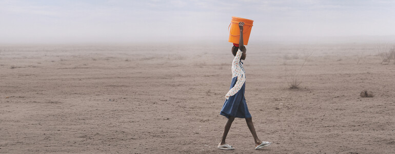 Person trägt orangefarbenen Wasserkanister durch ausgetrocknete Landschaft unter weitem Himmel, Thema Hunger und Wasserknappheit.