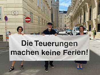 Drei Menschen halten ein Plakat mit der Aufschrift "Die Teuerungen machen keine Ferien!"