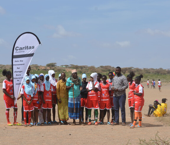 Eine Gruppe junger Frauen in roten Sporttrikots steht gemeinsam mit Erwachsenen und einem Banner am Spielfeldrand. Sie nehmen an einem Friedensprojekt teil, das Sport und Aufklärungsarbeit verbindet.