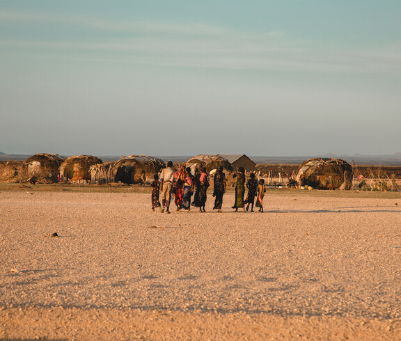 Eine Gruppe von Menschen geht über eine trockene, karge Ebene in Richtung traditioneller Rundhütten. Die Szene zeigt ein Nomadenlager in einer von Dürre betroffenen Region.