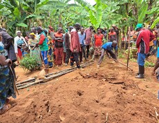 Eine größere Gruppe von Männern und Frauen steht aufmerksam im Freien zwischen Bananenstauden und beobachtet eine praktische Demonstration zur Bodenbearbeitung mit Holzwerkzeug auf rotem Lehmboden.