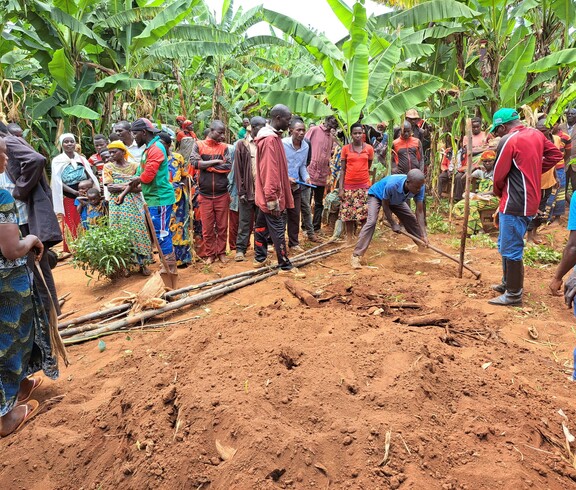 Eine größere Gruppe von Männern und Frauen steht aufmerksam im Freien zwischen Bananenstauden und beobachtet eine praktische Demonstration zur Bodenbearbeitung mit Holzwerkzeug auf rotem Lehmboden.