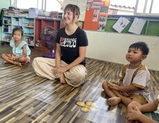 Eine junge Erwachsene sitzt mit mehreren Kindern im Kreis auf dem Boden eines Klassenzimmers.