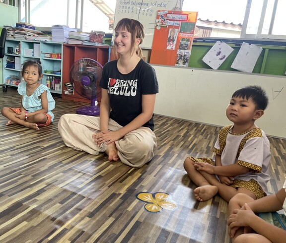 Eine junge Erwachsene sitzt mit mehreren Kindern im Kreis auf dem Boden eines Klassenzimmers.