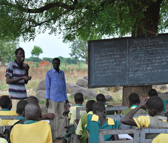 Schüler*innen und Lehrer beim Unterricht unter einem Baum, die Schultafel lehnt an den Stamm des Baumes.