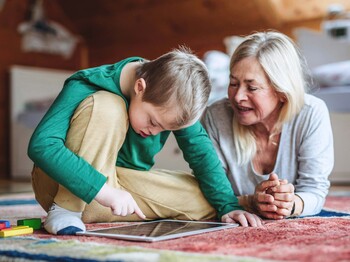 Kind sitzt auf einem bunten Teppich in einem Dachzimmer und nutzt ein Tablet, während eine erwachsene Person daneben liegt und aufmerksam zuschaut. Neben dem Tablet liegen bunte Spielsteine, im Hintergrund ein Bett und Holzdecke.