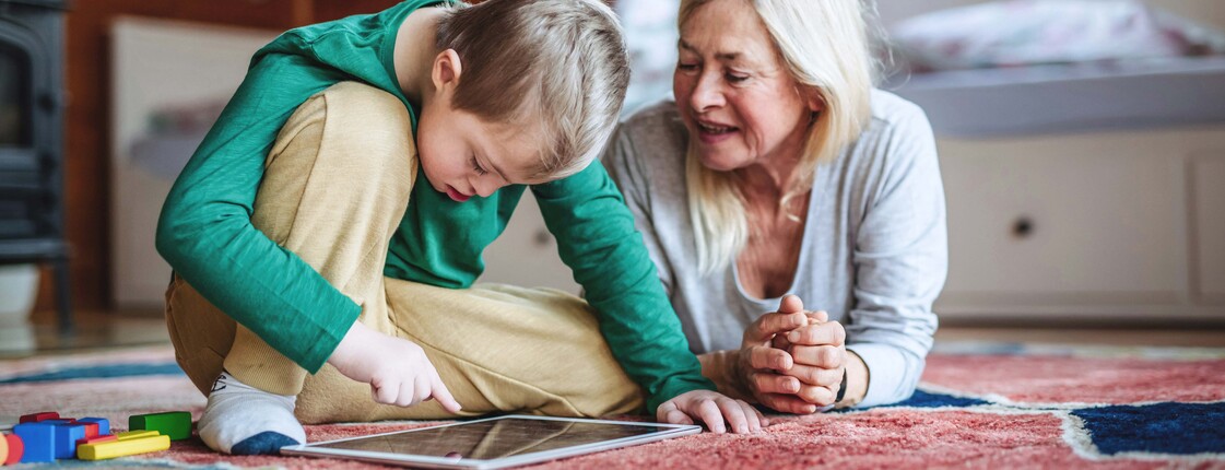 Kind sitzt auf einem bunten Teppich in einem Dachzimmer und nutzt ein Tablet, während eine erwachsene Person daneben liegt und aufmerksam zuschaut. Neben dem Tablet liegen bunte Spielsteine, im Hintergrund ein Bett und Holzdecke.