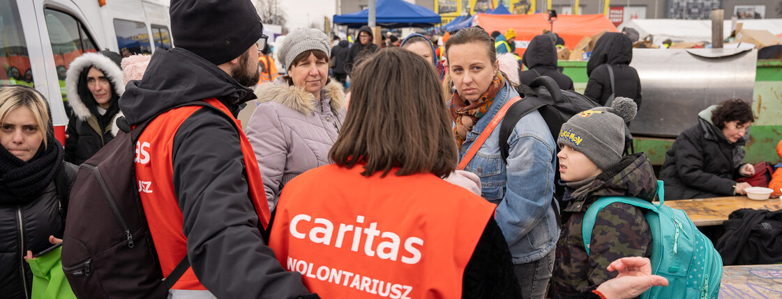 Man sieht zwei Freiwillige der Caritas von hinten, sie tragen rote Westen mit Caritas-Logo, sie sprechen mit ankommenden Geflüchteten aus der Ukraine