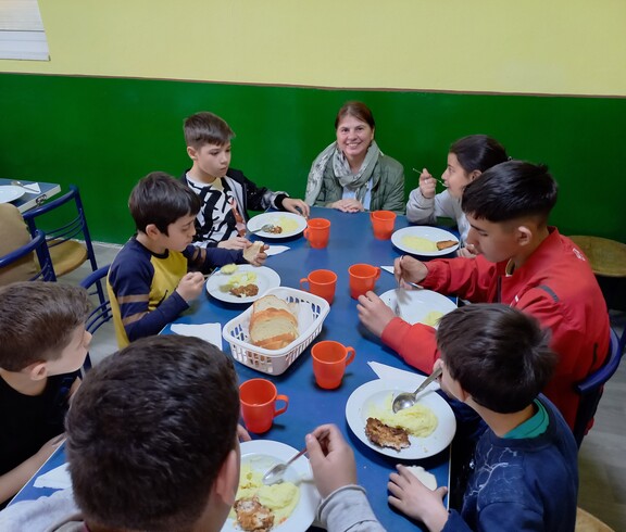 Eine Gruppe Kinder sitzt am Tisch und isst. Eine Frau kniet daneben. 