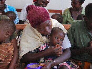Eine Mutter mit Kopftuch füttert ihr kleines Kind behutsam mit der Hand; links daneben sitzt ein weiteres Kleinkind – Szene aus dem Ernährungszentrum im Waisenhaus Gitega, Burundi.