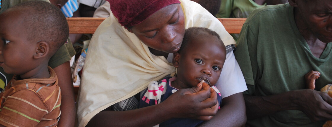 Eine Mutter mit Kopftuch füttert ihr kleines Kind behutsam mit der Hand; links daneben sitzt ein weiteres Kleinkind – Szene aus dem Ernährungszentrum im Waisenhaus Gitega, Burundi.