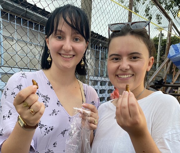 Zwei junge Erwachsene halten regionale Snacks in der Hand und lächeln in die Kamera.