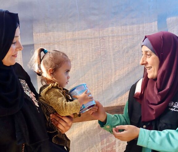 Eine Mitarbeiterin der Caritas Jerusalem überreicht einer Frau mit Kind eine Dose Babynahrung. Beide Frauen lächeln sich an.