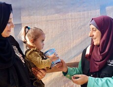 Eine Mitarbeiterin der Caritas Jerusalem überreicht einer Frau mit Kind eine Dose Babynahrung. Beide Frauen lächeln sich an.