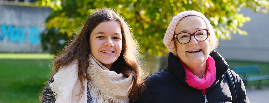 Besuchs- und Begleitdienste Eine junge Frau begleitet eine Seniorin, beide tragen Winterkleidung, beim Spazierengehen durch einen Park.