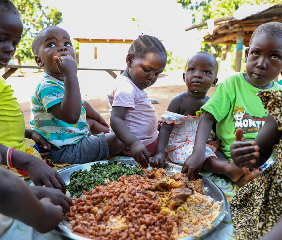 Kinder sitzen im Freien um eine große Schüssel mit Reis, Bohnen und Gemüse und essen gemeinsam mit den Händen.