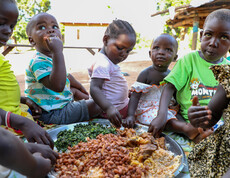 Kinder sitzen im Freien um eine große Schüssel mit Reis, Bohnen und Gemüse und essen gemeinsam mit den Händen.