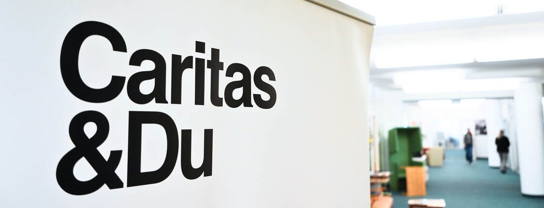 Schriftzug "Caritas & Du" auf Flipchart, im Hintergrund ist verschwommen ein Büro zu erkennen