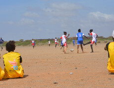 Mehrere Afrikaner*innen spielen auf einem sandigen Platz Fußball. Zwei Teams in unterschiedlichen Trikots treten gegeneinander an, während eine weitere Person am Spielfeldrand sitzt und zuschaut.