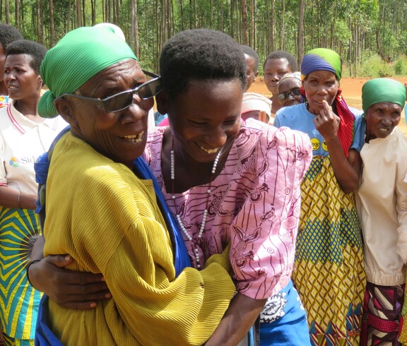 zwei burundische Frauen lachen und umarmen sich. Im Hintergrund sieht man weitere Frauen stehen. 