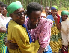 zwei burundische Frauen lachen und umarmen sich. Im Hintergrund sieht man weitere Frauen stehen. 