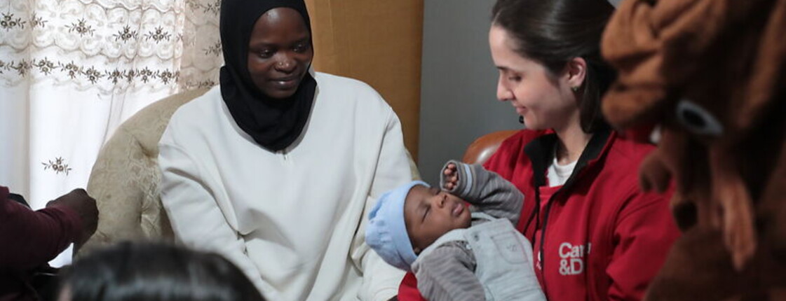Eine Frau mit roter Caritas-Jacke hält ein Baby in Händen. Die Mutter des Babys sitzt daneben.