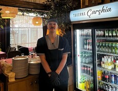 Junger Mann in Arbeitskleidung steht in einem Restaurant neben einem Kühlschrank mit Getränken und gestapeltem Geschirr.