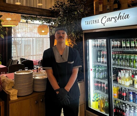 Junger Mann in Arbeitskleidung steht in einem Restaurant neben einem Kühlschrank mit Getränken und gestapeltem Geschirr.